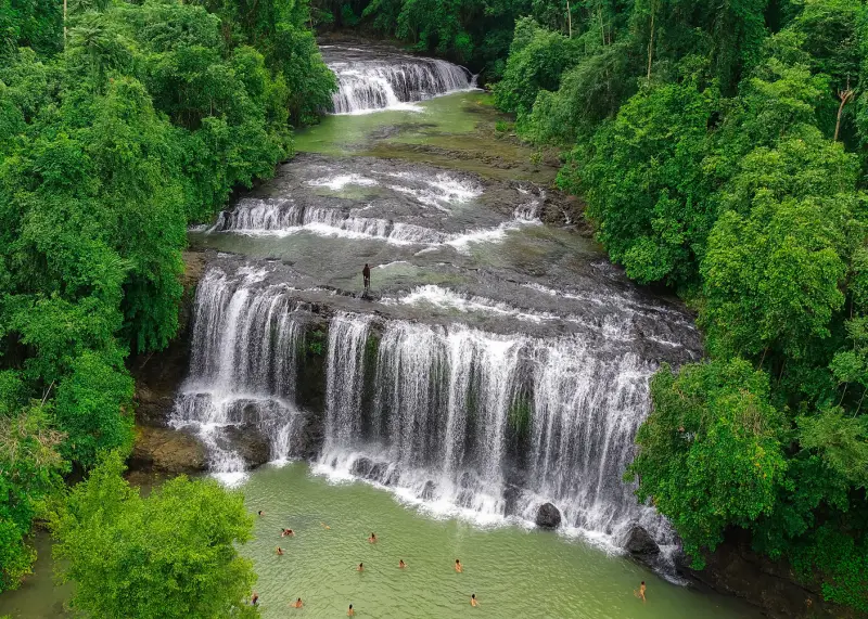 Multi-tier waterfall in the Philippine jungle with locals standing on rocks and lush greenery surrounding.
