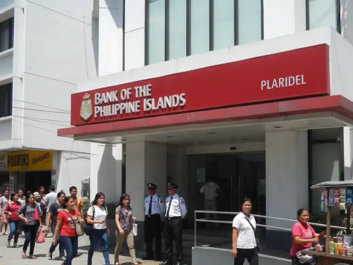 A candid Filipino street scene shows a BPI Plaridel branch with two security guards in white uniforms, a street vendor selling cigarettes and candies, and many people walking by.