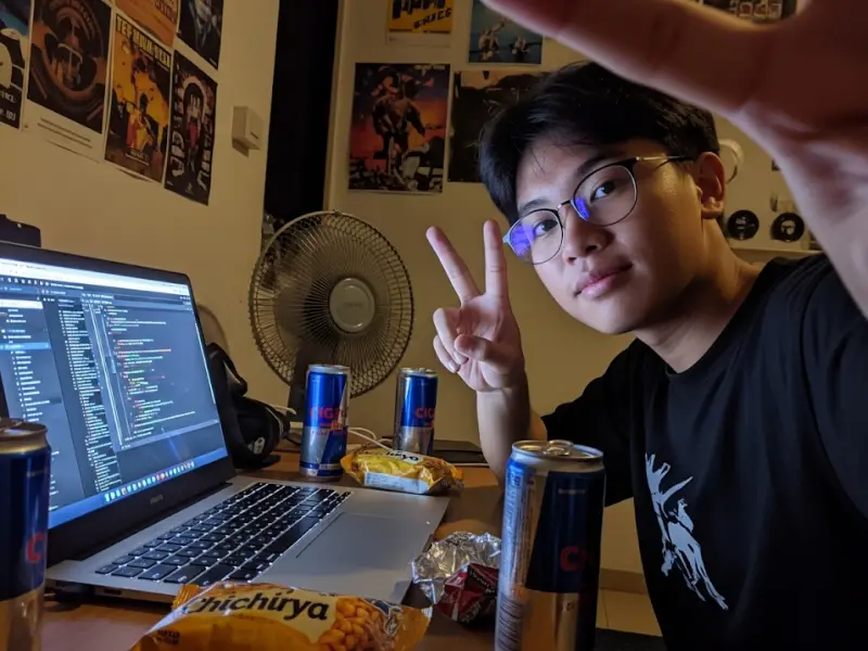Web developer selfie coding on laptop at night with snacks and fan nearby.