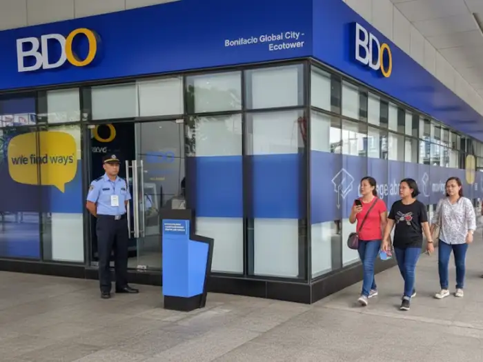 A candid shot of a busy Filipino street scene with people walking on the sidewalk outside a BDO bank branch, while a security guard stands near the entrance.